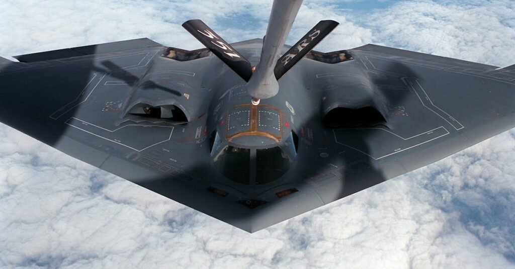 A B-2 Stealth Bomber aircraft receives aerial refueling mid-flight over clouds.
